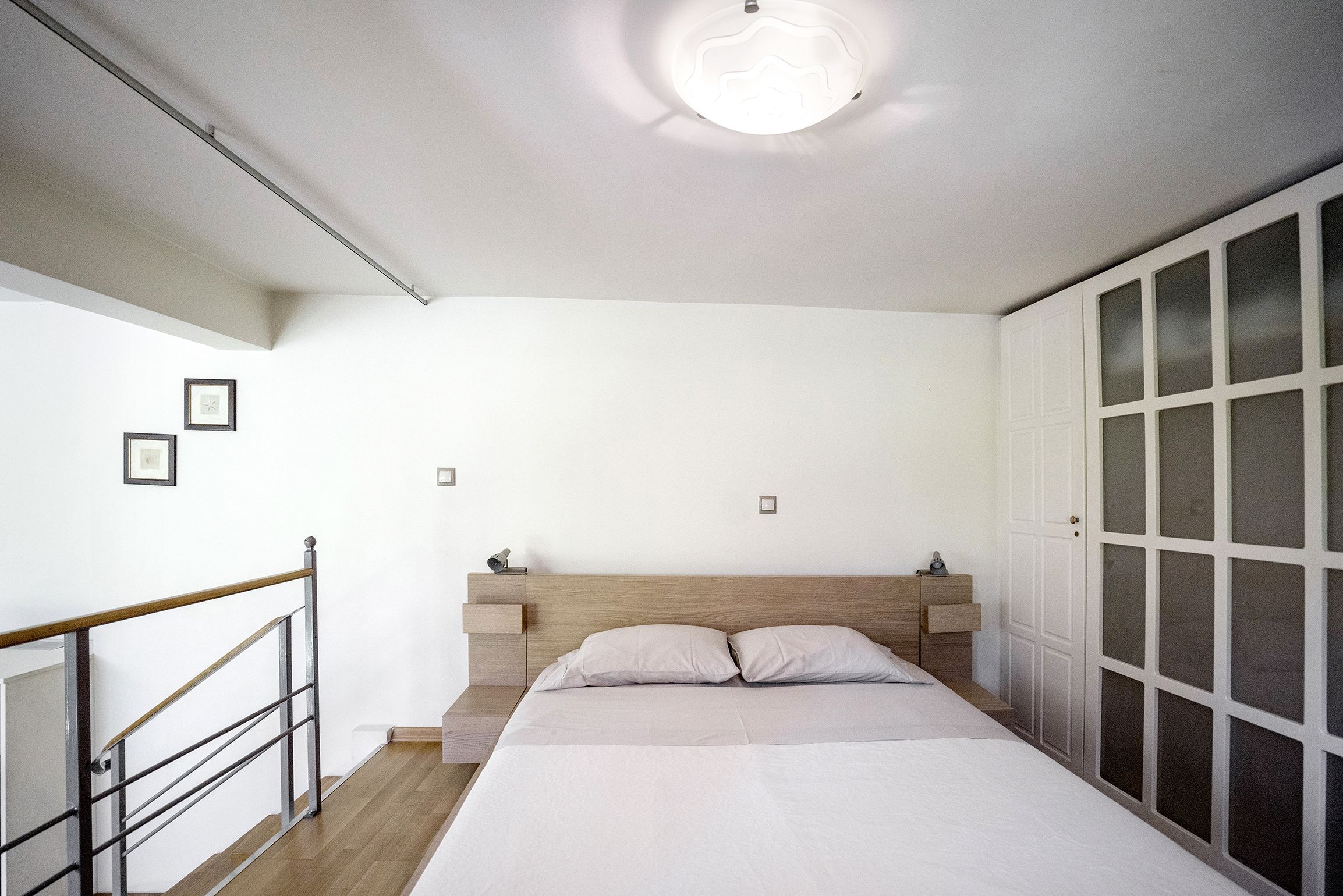A bedroom with a wooden headboard, a bed with two pillows, a nightstand, and a wardrobe with frosted glass doors.