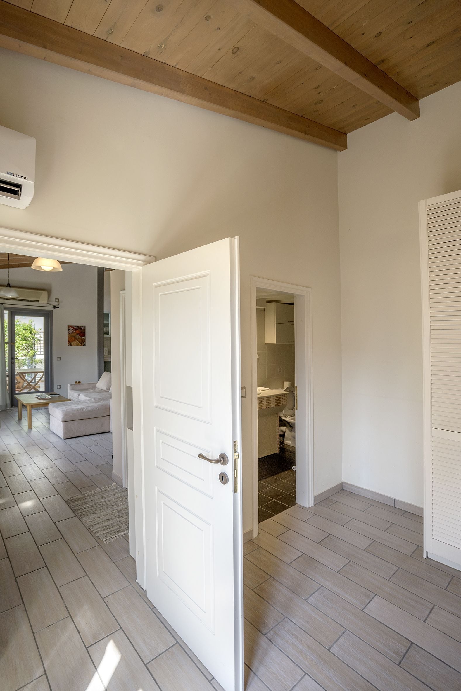 A hallway with a white door leading to a bathroom, a living area with a sofa and a table, and a ceiling with wooden beams.