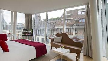 Una habitación de hotel con cama, sofá, televisión y ventanas amplias con vista a la ciudad.