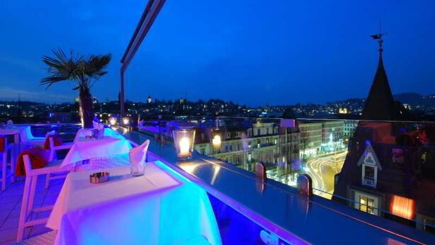 A rooftop bar with tables and chairs, a palm tree, and a cityscape view.
