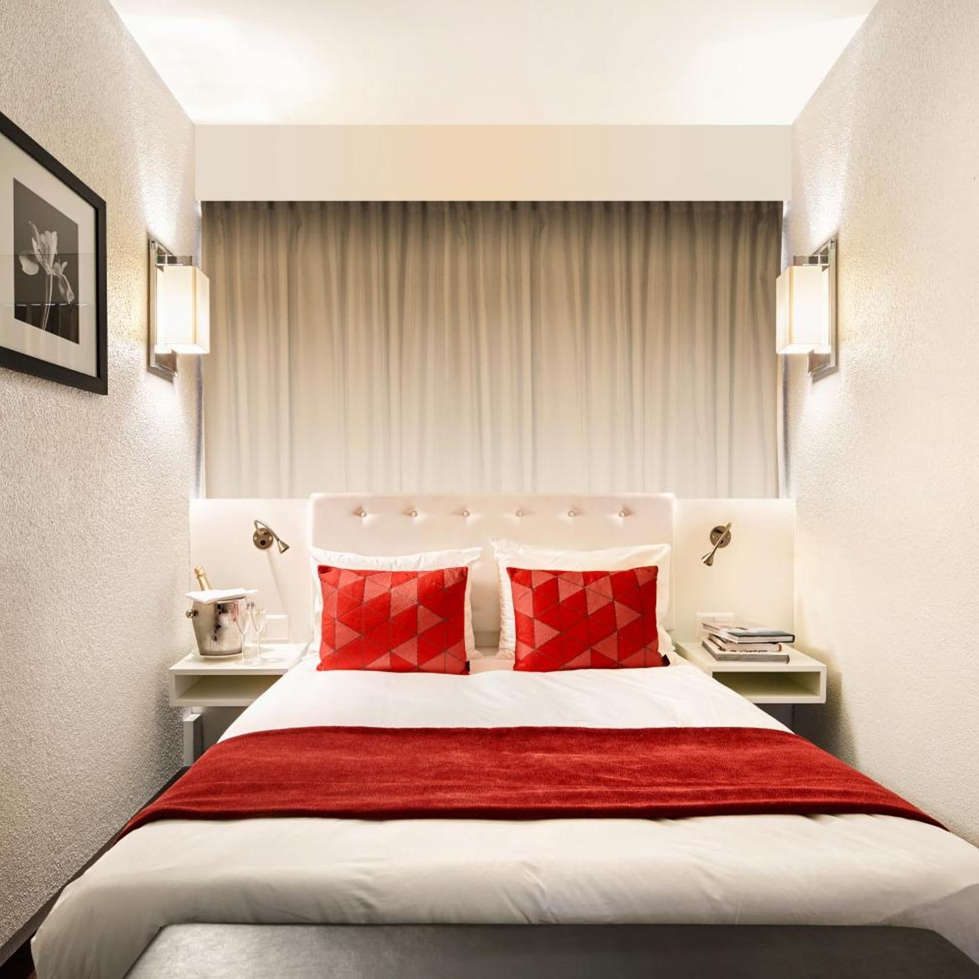 Una habitación de hotel con una cama, dos mesitas de noche, un cojín rojo y blanco estampado, una manta roja y un cuadro en la pared.
