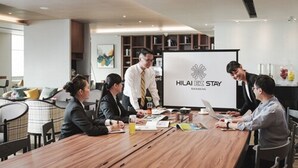 Un salón de conferencias con una pizarra mostrando el logotipo de Hilai ZZ Stay, una mesa de madera y participantes reunidos en una reunión.