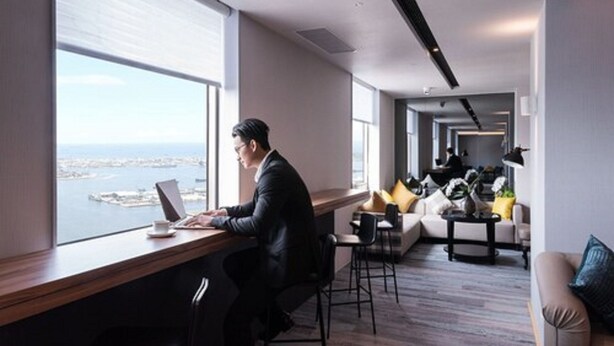 Un espacio de oficina moderno con vista al mar, un escritorio con una computadora portátil y una zona de estar con sofás y una mesa de centro.