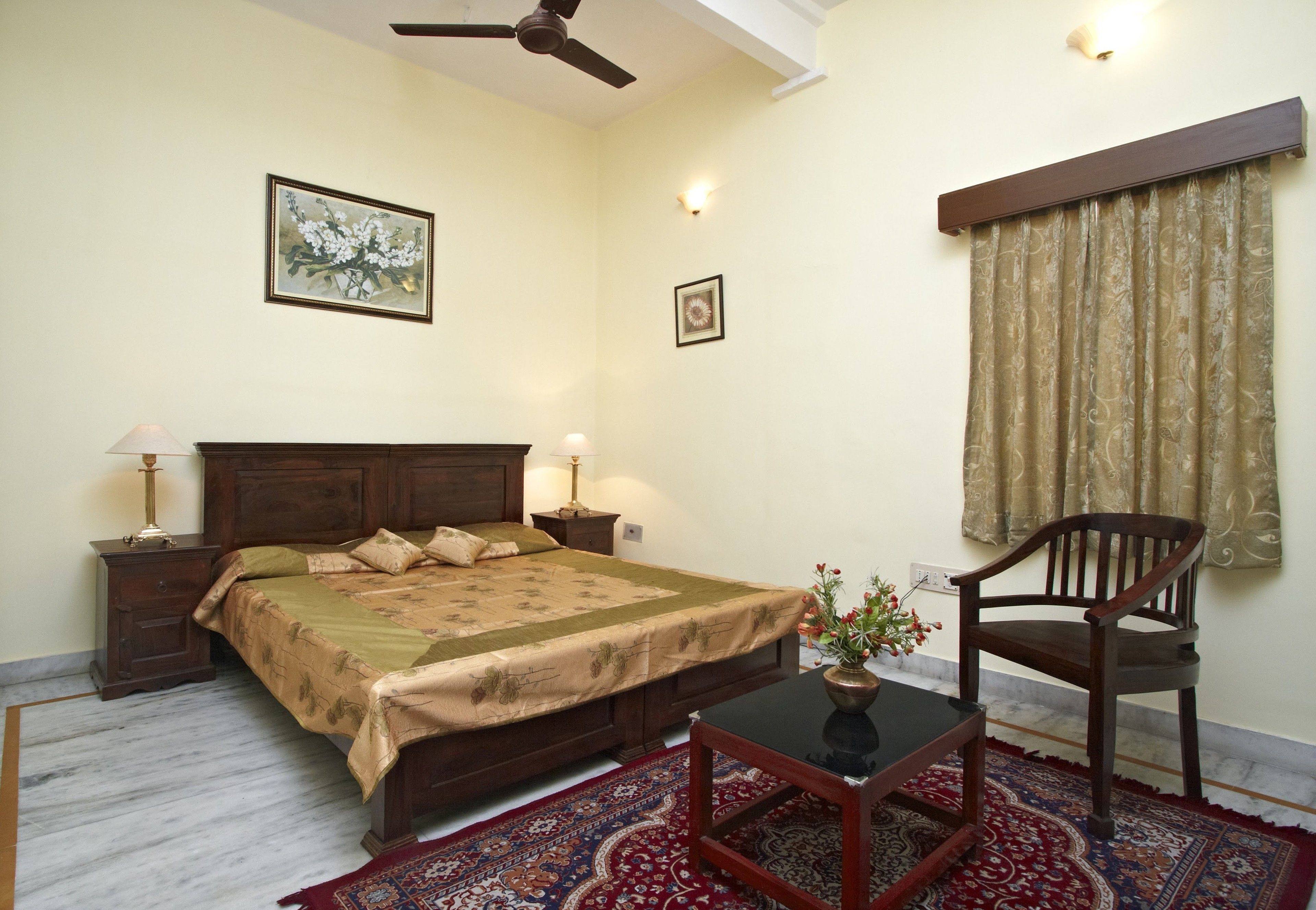 A bedroom with a bed, bedside tables, a chair, a table, a ceiling fan, and a window with curtains.