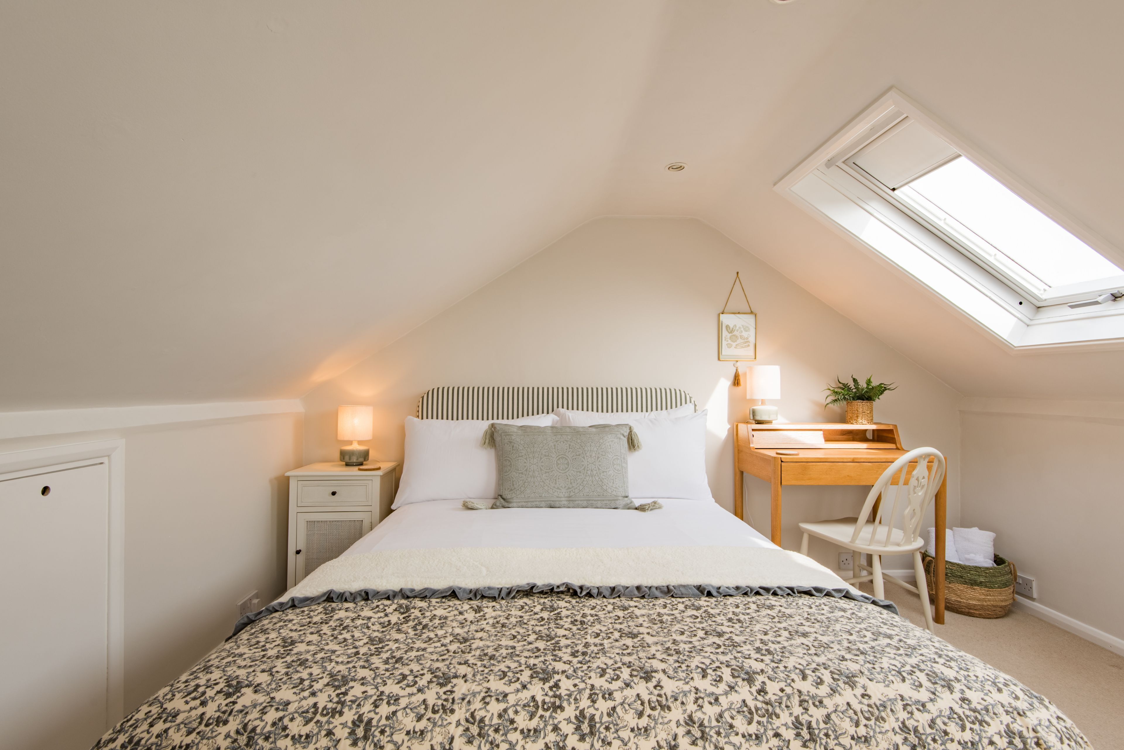 A bedroom with a sloped ceiling, a skylight, a bed with a patterned bedspread, a bedside table with a lamp, a desk with a chair, and a small plant.