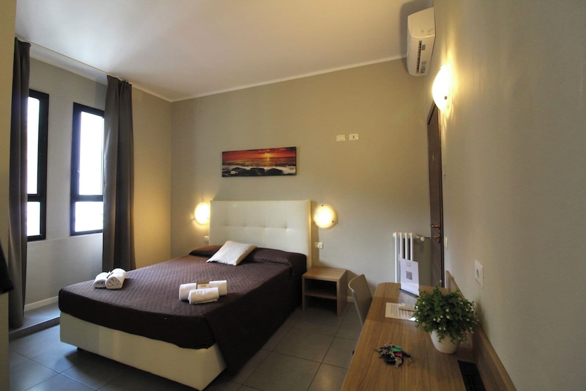 A hotel room with a bed, bedside tables, a painting, a potted plant, and a window.