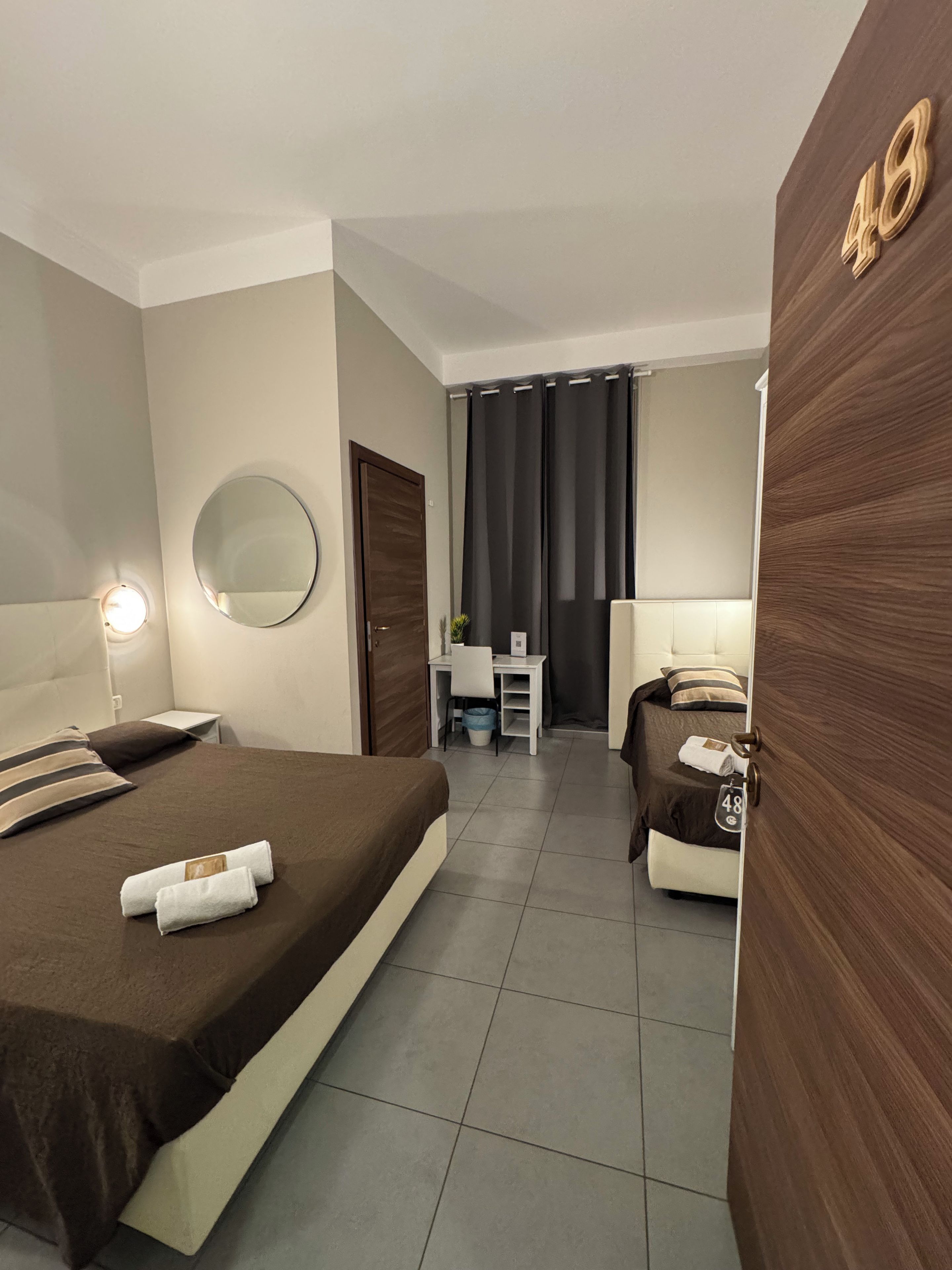 A hotel room with a bed, a desk, a chair, a mirror, and a door with a number.