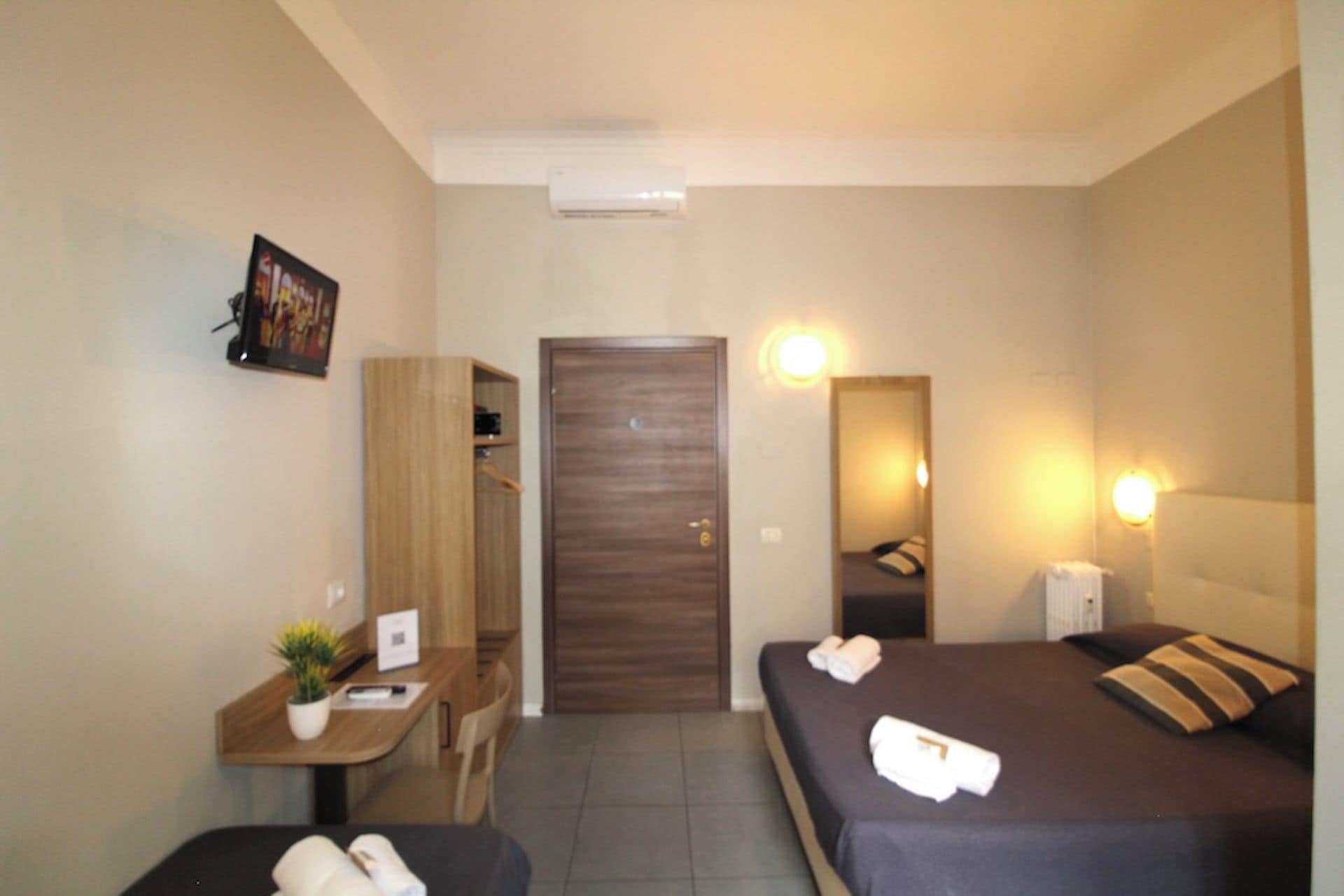 Una camera d'albergo con un letto, un comodino, una scrivania, una sedia, un armadio, una porta e un condizionatore d'aria a parete.