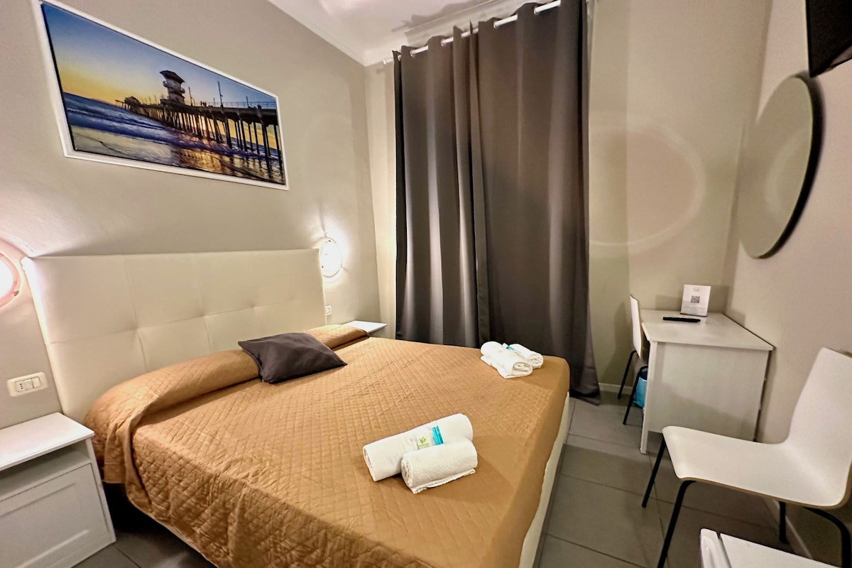 A hotel room with a bed, a desk, a chair, a painting, and a curtain.