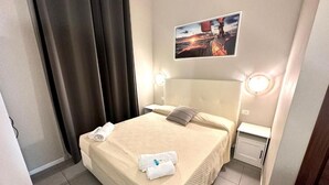 A neatly made bed with a beige cover, a pillow, and two rolled towels. There is a small bedside table with a lamp and a wall-mounted picture. - Hotel Golden Milano (Milan)