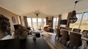 Un amplio salón con un gran ventanal que ofrece vistas panorámicas, un comedor con mesa y sillas de madera, y una cocina con encimera negra y armarios blancos.