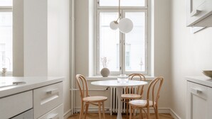 A modern kitchen with white cabinetry, a round dining table with four wooden chairs, a hanging light fixture, and a window with blinds. - Roost Roobert (Helsinki)