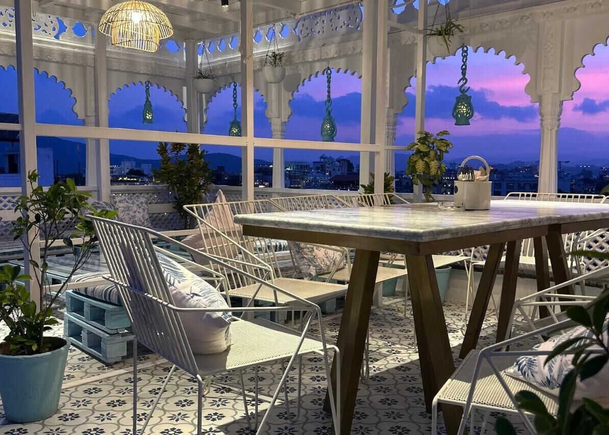Un espace extérieur aménagé avec une table et des chaises, entouré de plantes en pot et offrant une vue sur le paysage urbain au crépuscule.