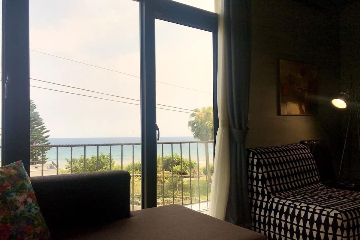 A room with a sofa and a patterned chair, a large window with a view of the sea, and a painting on the wall.