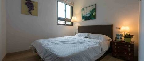 Un dormitorio con una cama, una mesita de noche con lámpara, una ventana y dos cuadros en la pared.