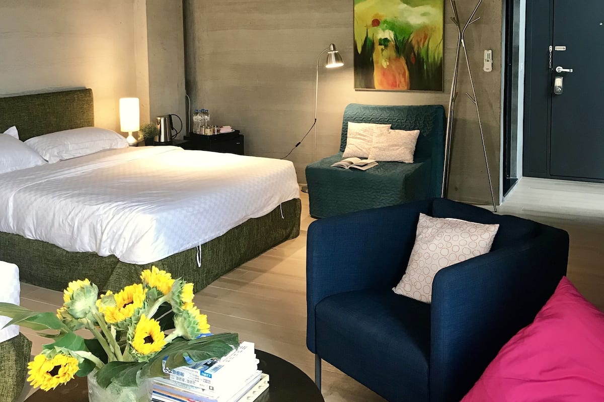 A modern hotel room with a large bed, a sofa, a coffee table with books and flowers, and a painting on the wall.