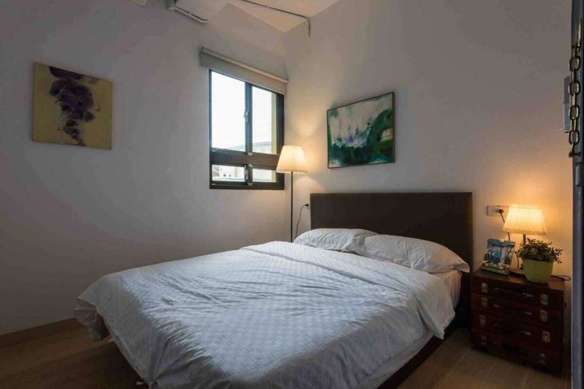 A bedroom with a bed, a nightstand with a lamp, a window, and two paintings on the wall.