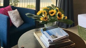 Een kamer met uitzicht op zee, een blauwe bank met een gestreept kussen, een tafel met een boek en een vaas vol zonnebloemen.