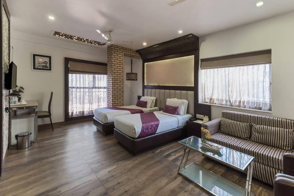 Habitación de hotel con dos camas, un sofá, una mesa de centro de vidrio, un escritorio y un cesto de basura.