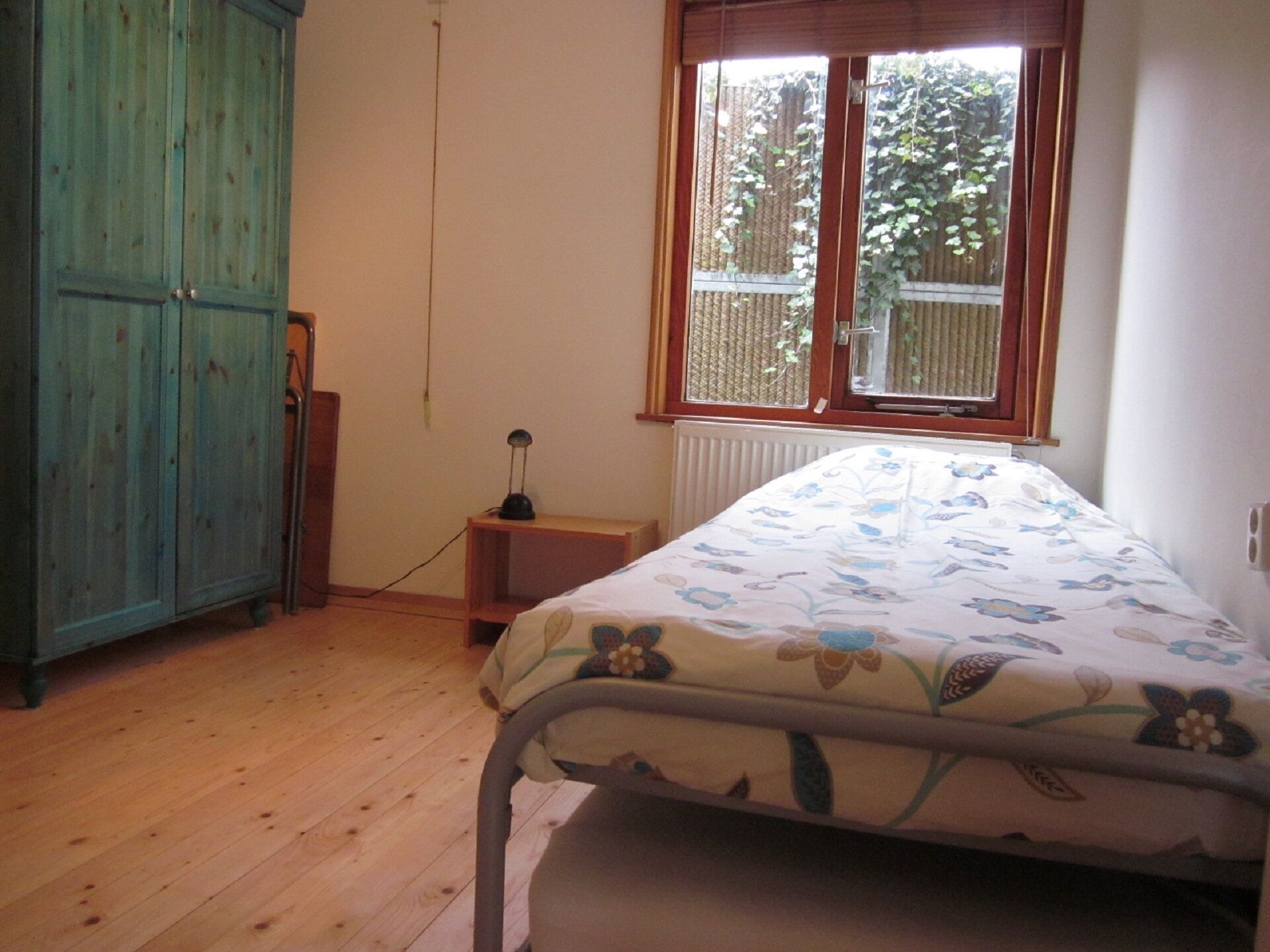 A bedroom with a wooden floor, a bed with floral bedding, a window with a view of greenery, and a teal wardrobe.