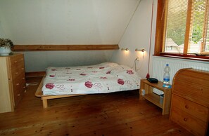 Une petite chambre avec un lit simple, un plancher en bois, une table de chevet, une commode et une fenêtre avec des stores.