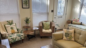 A cozy living room with a sofa, armchair, and coffee table. There are potted plants and a framed picture on the walls.