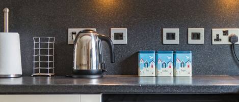 A kitchen with a kettle, tea, coffee, and sugar boxes, a washing machine, and a power outlet.