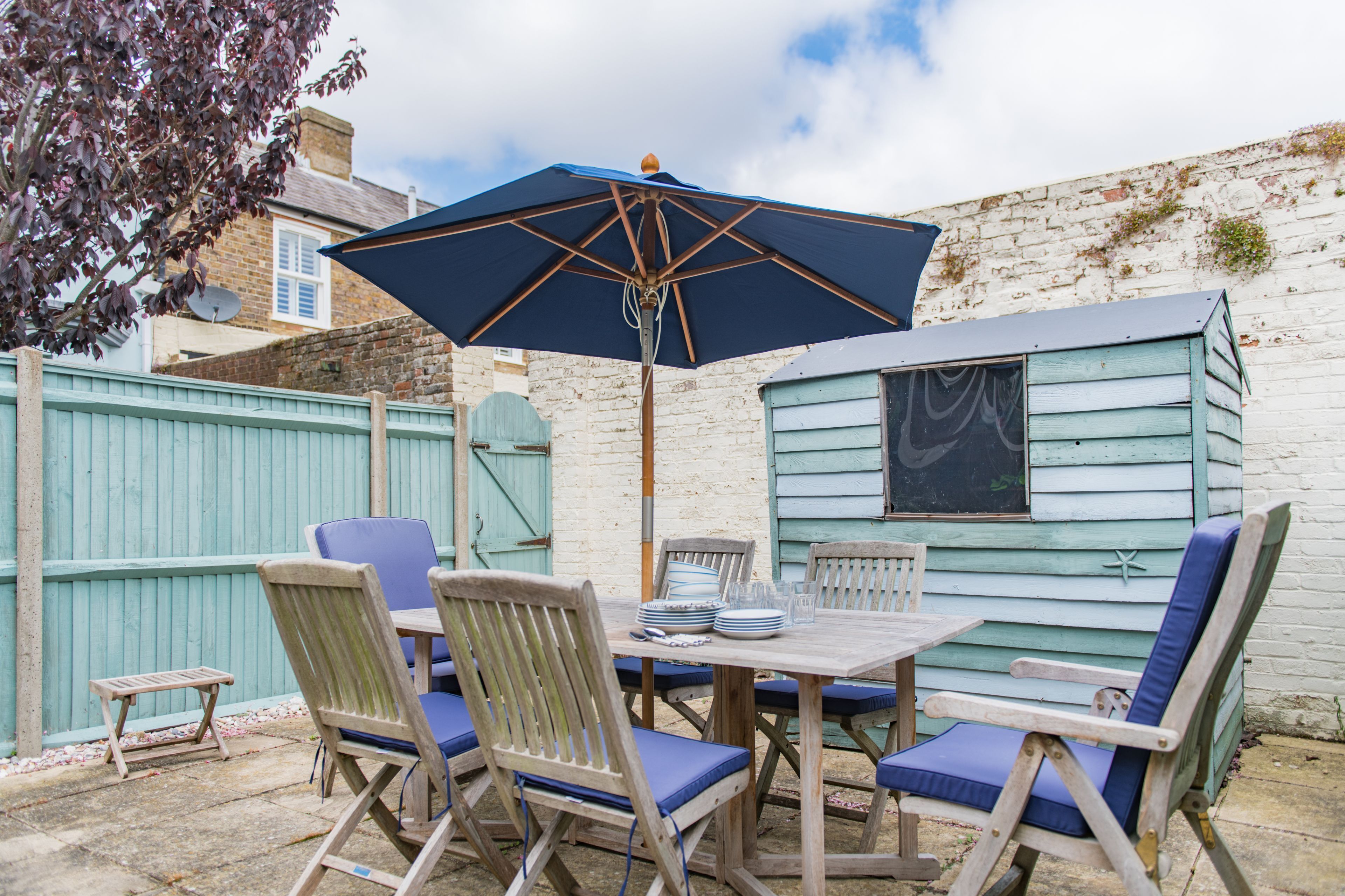 A wooden table with chairs and a blue umbrella in a backyard with a brick wall and a shed.