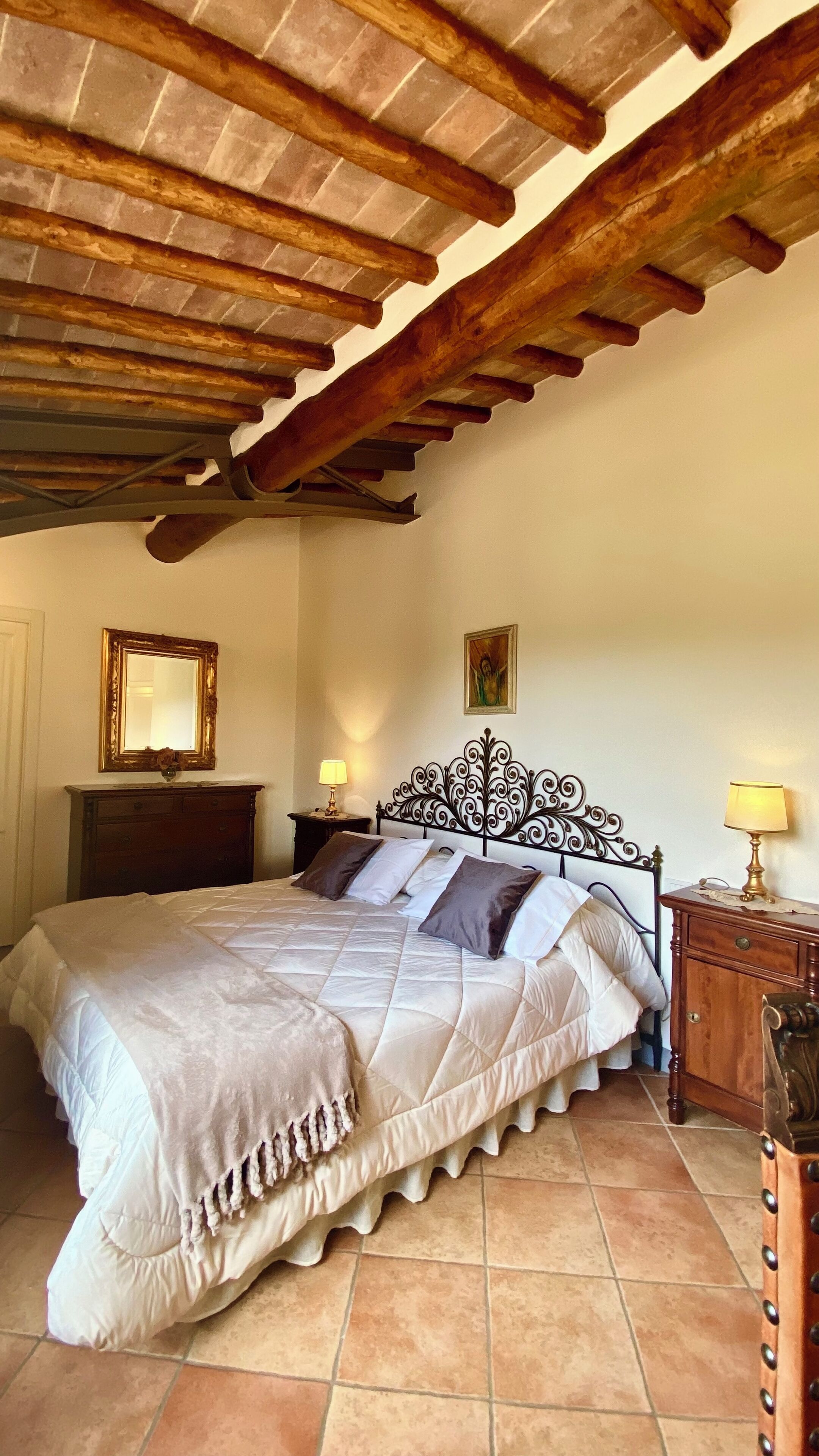 A bedroom with a large bed, wooden ceiling beams, a dresser, a mirror, and a picture on the wall.
