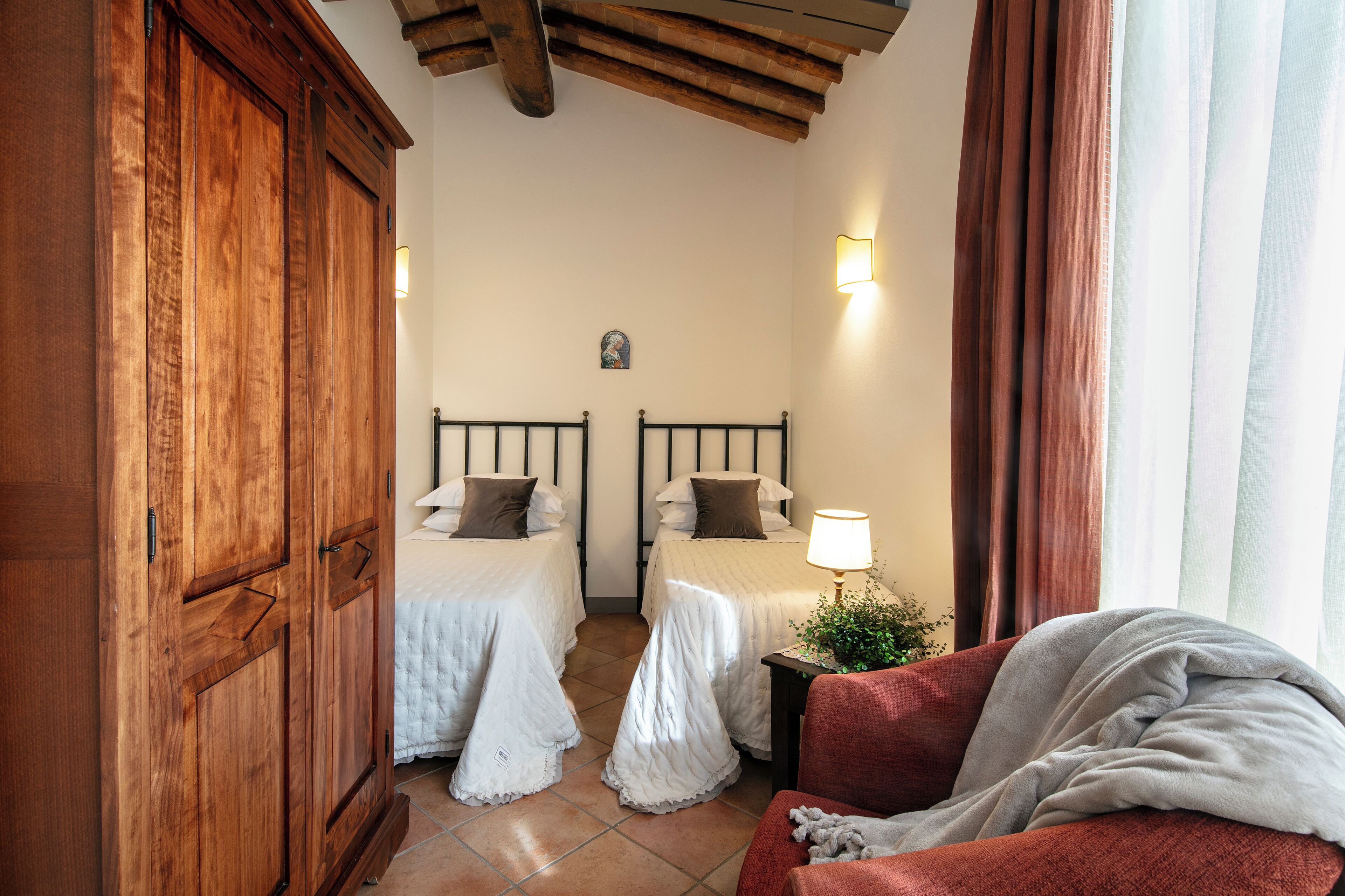 Habitación de hotel con dos camas, un armario de madera, un sillón rojo y una ventana con cortinas.