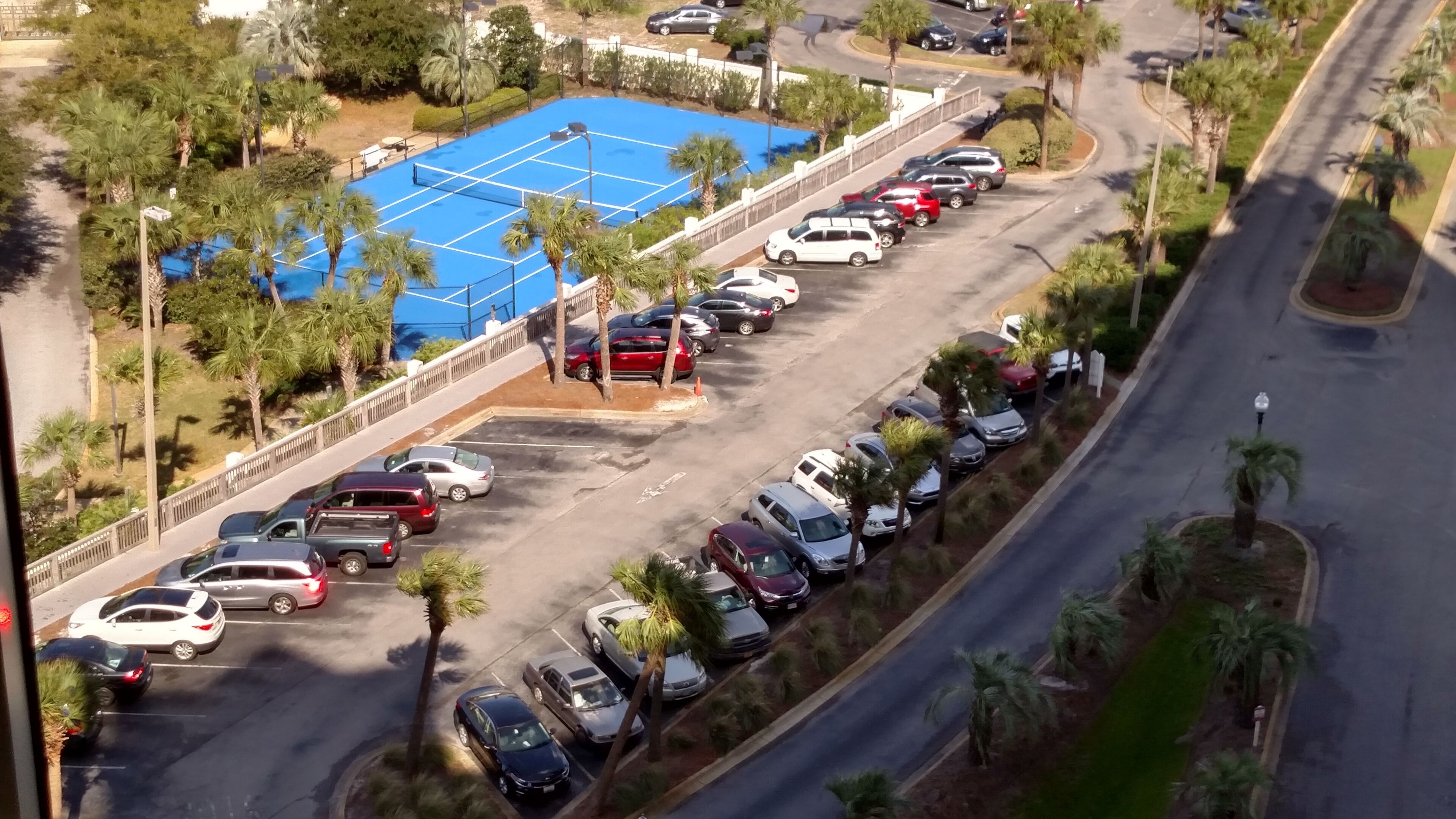 Vista aérea de un estacionamiento con varias filas de coches aparcados, una cancha de tenis azul y palmeras.