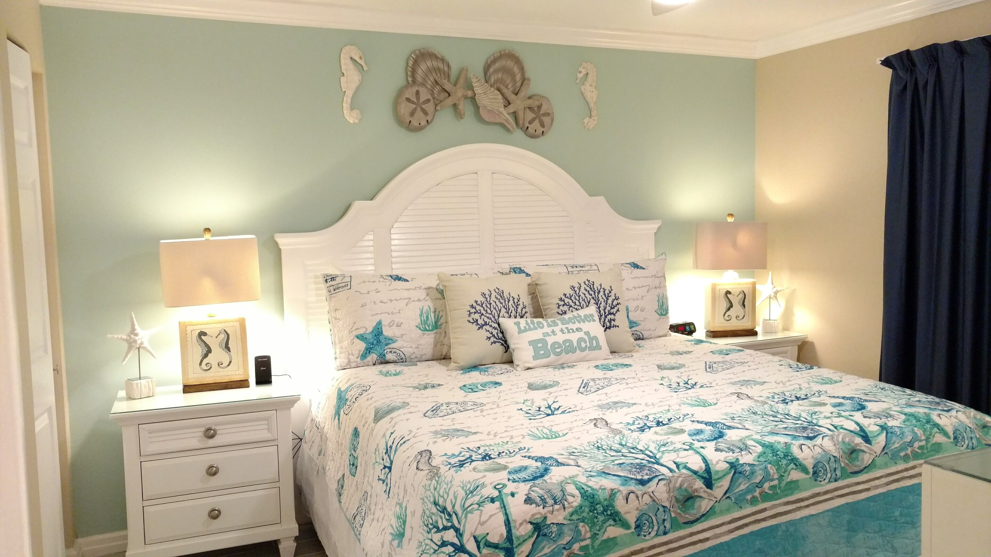 Un dormitorio con cama blanca, varios almohadones, una mesita de noche, una lámpara y adornos en la pared.