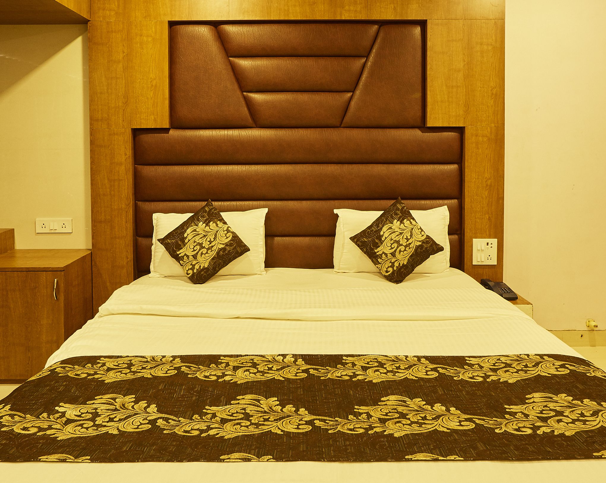 Habitación de hotel con una cama grande, un cabecero de madera, dos almohadones decorativos y una mesita de noche con un teléfono.
