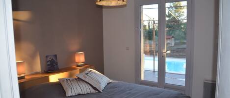 A bedroom with a bed, two bedside lamps, a painting, and a view of a swimming pool through a glass door.