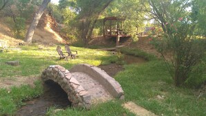 A rustic stone bridge over a small stream, surrounded by lush greenery and a wooden gazebo.