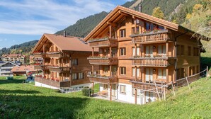 A wooden chalet-style building with multiple balconies, surrounded by lush greenery and mountains.