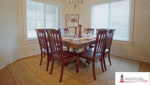 A dining room with a wooden table set for six, surrounded by six wooden chairs. There is a chandelier above the table and a framed picture on the wall.
