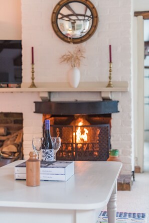 Una chimenea encendida, una mesa blanca con una botella y una copa, y un espejo sobre la repisa.