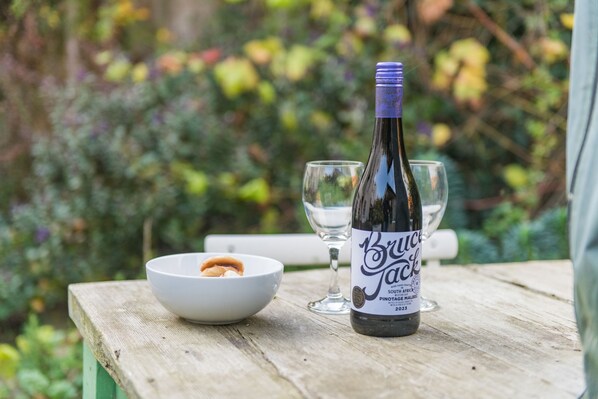 Una botella de Bruce Jackson Pinotage sobre una mesa de madera, junto a un recipiente con comida y dos copas.