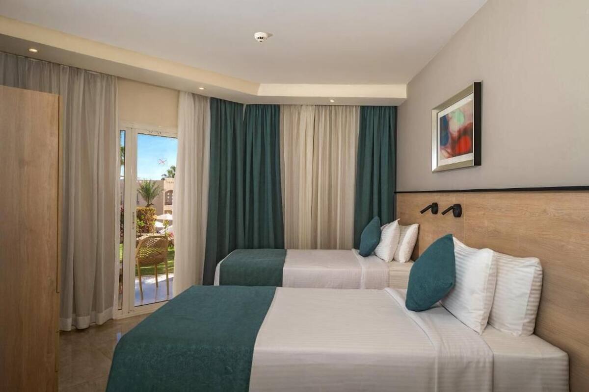 A hotel room with two beds, a large window with curtains, a view of the outdoors, and a painting on the wall.