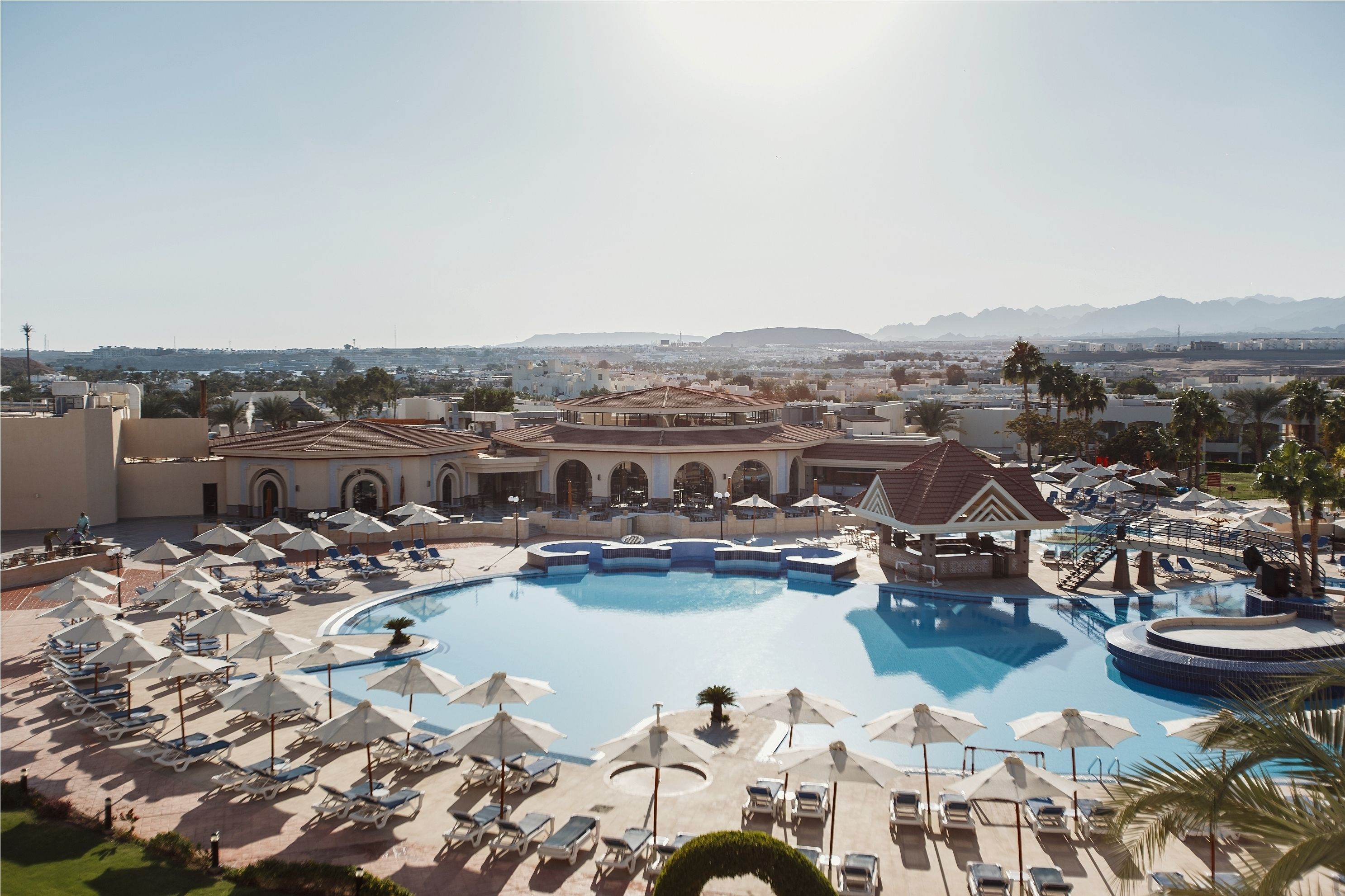 Ein Resort mit großem Pool, Liegestühlen, Sonnenschirmen und Blick auf ferne Berge.