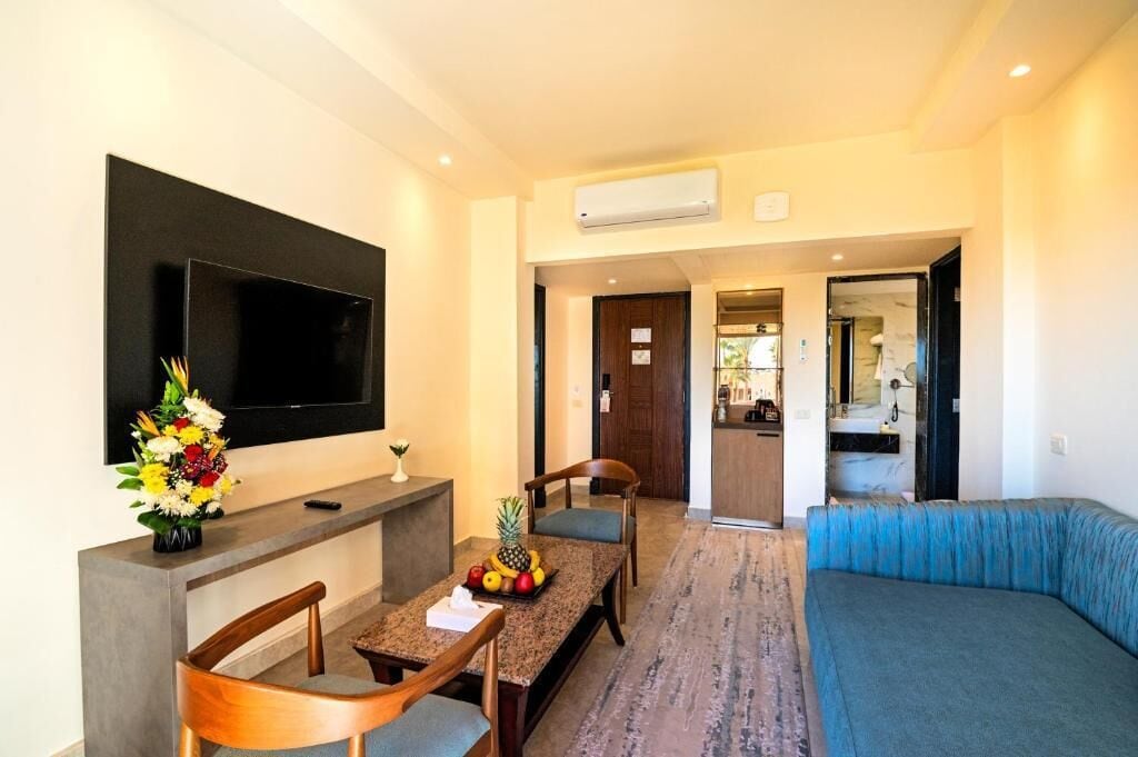 A modern hotel room with a flat-screen TV, a dining area with a fruit basket, and a blue sofa.