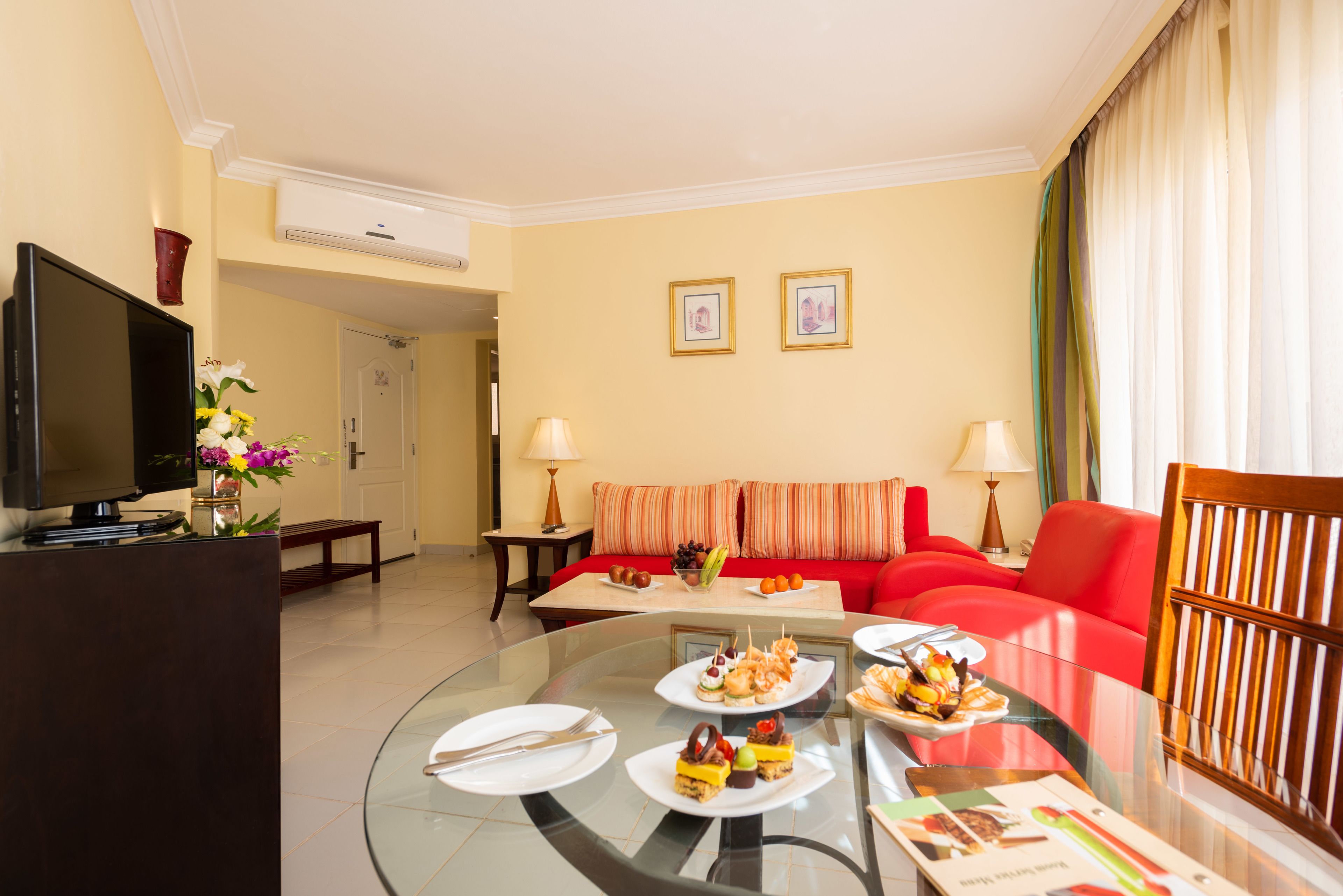 Habitación de hotel con una mesa de vidrio dispuesta para una comida, un sofá rojo, un televisor y cuadros en la pared.
