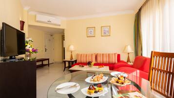 Habitación de hotel con una mesa de vidrio dispuesta para una comida, un sofá rojo, un televisor y cuadros en la pared.
