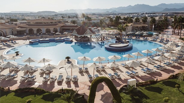A large pool surrounded by sun loungers and umbrellas, with a view of mountains in the background.