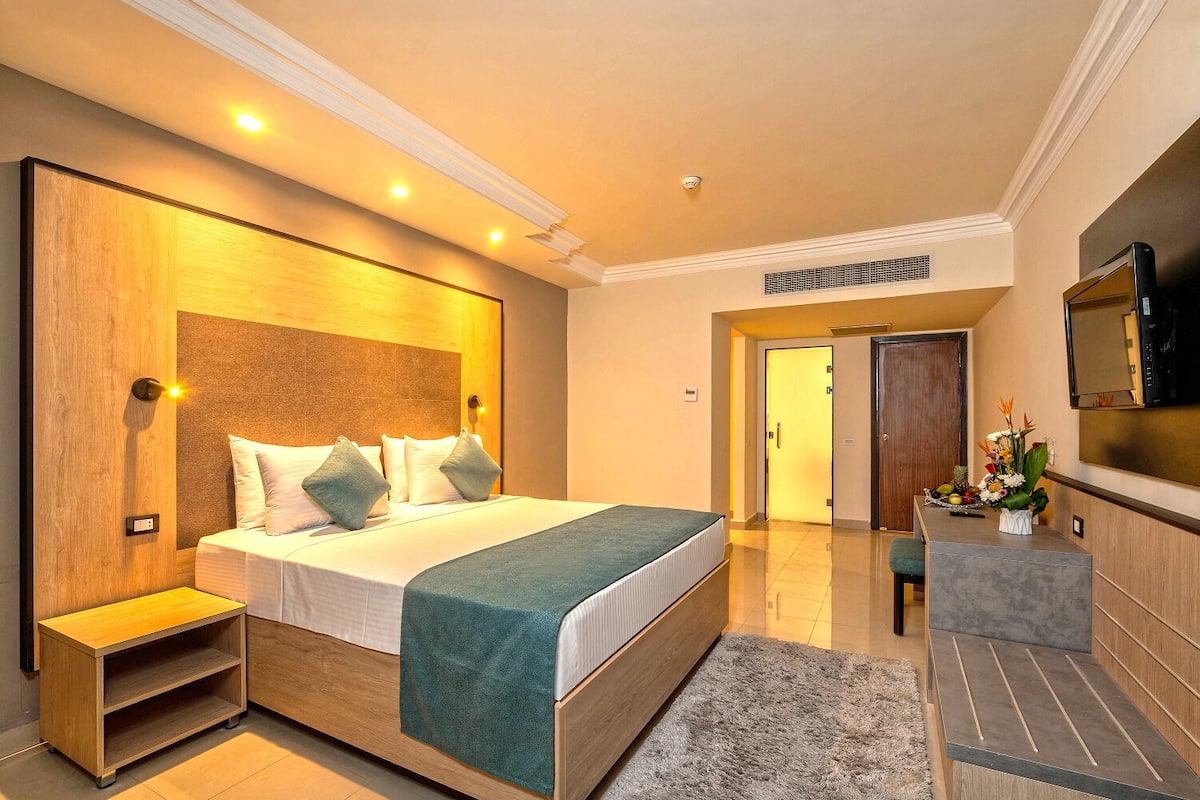 A modern hotel room with a large bed, bedside tables, a TV, and a bathroom visible through an open door.