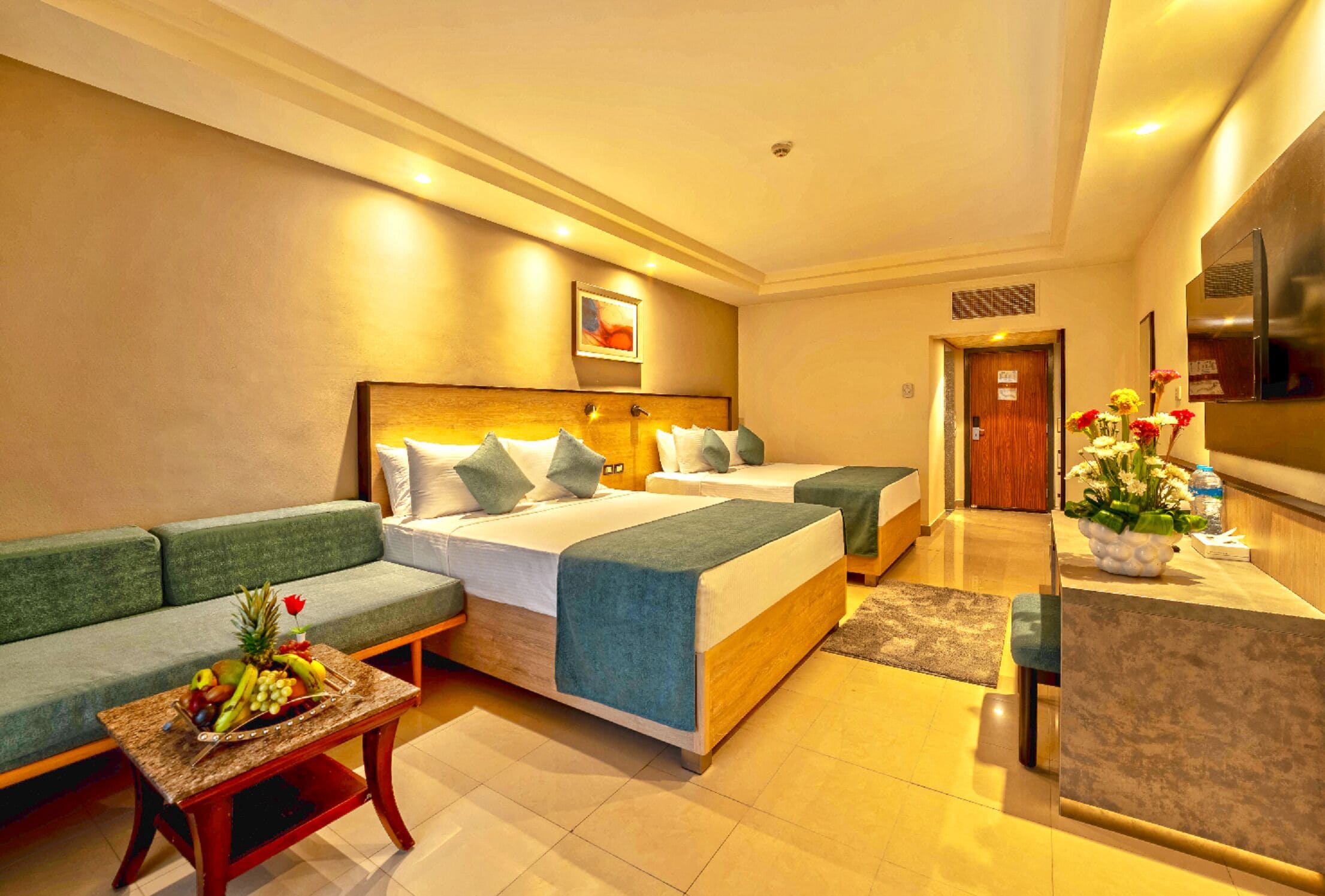 A hotel room with two beds, a sofa, a small table with fruit, and a kitchen area.