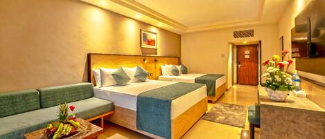 A hotel room with two beds, a sofa, a small table with fruit, and a kitchen area.