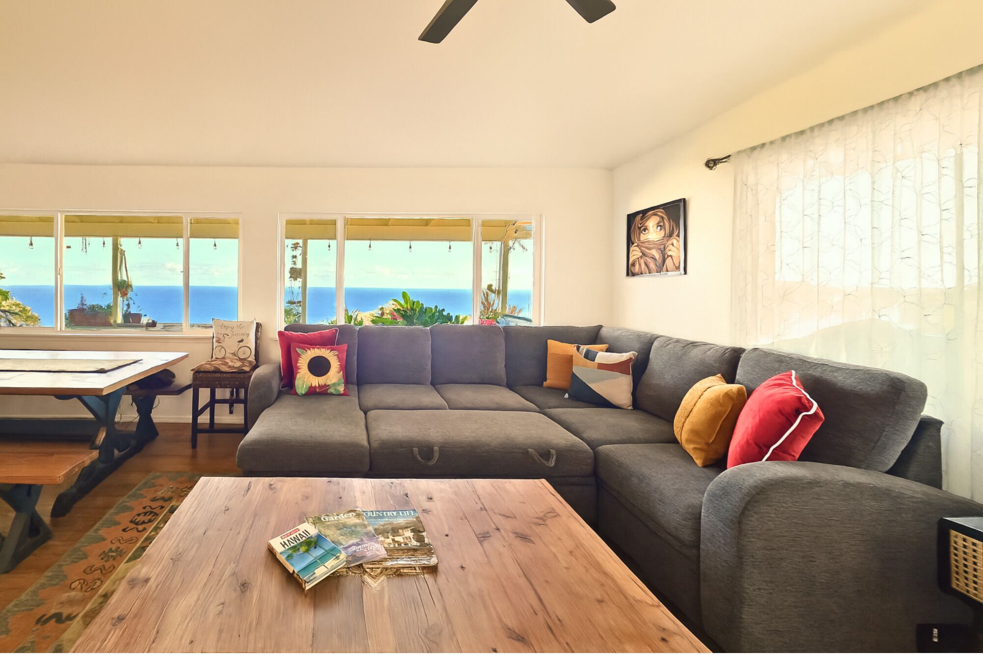 Ein Wohnzimmer mit einem grauen Ecksofa, einem Couchtisch mit Zeitschriften und Meerblick durch große Fenster.