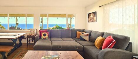 Ein Wohnzimmer mit einem grauen Ecksofa, einem Couchtisch mit Zeitschriften und Meerblick durch große Fenster.